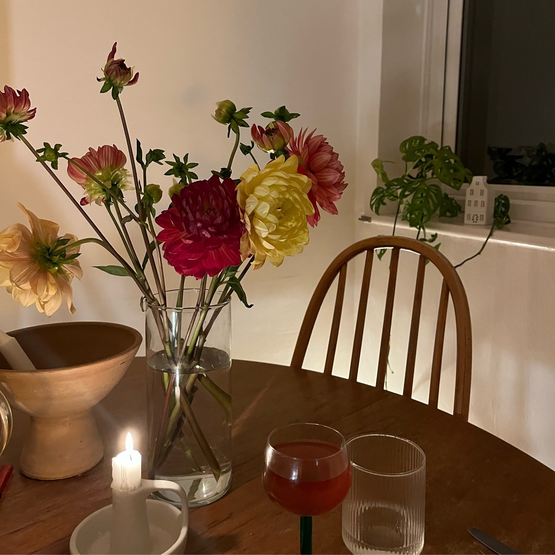 Dahlias on a vase on top of a brown table next to a candle burning and a glass of red wine.